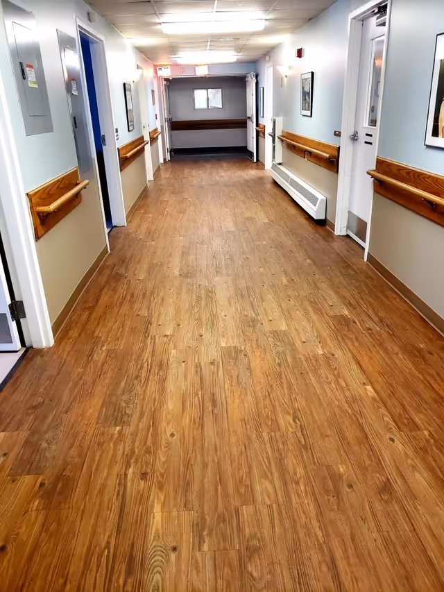 A long, clean hallway with wood-patterned flooring and light blue walls. The hallway has wooden handrails on both sides, several doors, framed pictures on the walls, and ceiling lights illuminating the space.