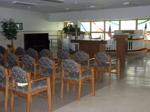 Rows of wooden chairs with patterned cushions face a stage in a small interior hall.
