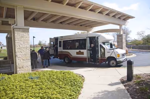 A shuttle bus is parked at the covered entrance of Ovation Sarah Chudnow, with residents boarding.
