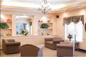 A cozy living room area with four cushioned armchairs arranged around a central space. The room features a large mirror on the back wall, decorative shelves with plants and ornaments, a chandelier hanging from the ceiling, and a window with draped curtains letting in natural light. There are also flower arrangements on the shelves and near the window.