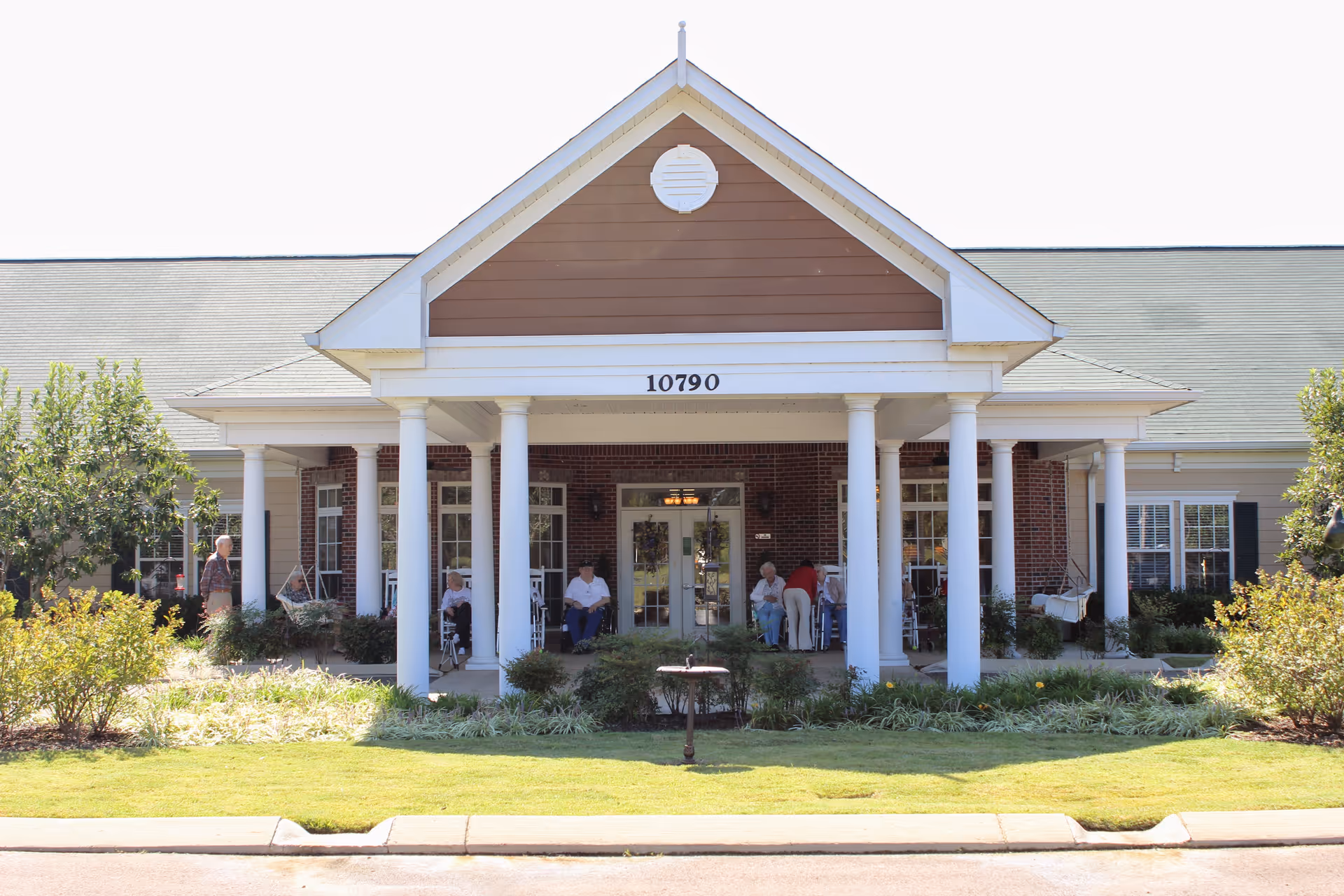 Front exterior view of a senior living facility with a covered porch supported by white columns. Several elderly people are sitting on rocking chairs on the porch. The building has a brick facade with white trim and the number 10790 displayed above the entrance. There are bushes and a well-maintained lawn in front of the building.
