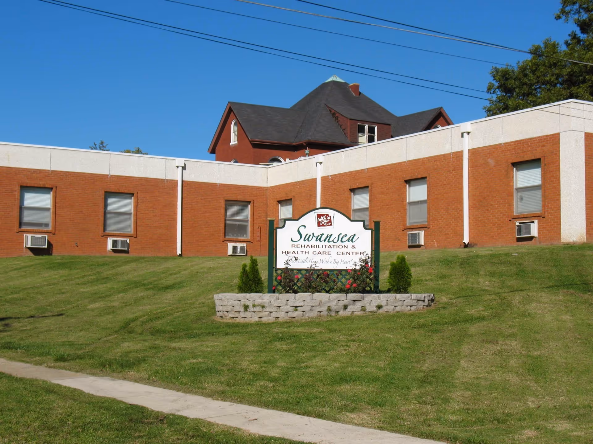 Exterior view of Swansea Rehabilitation & Health Care Center, a brick building with multiple windows and air conditioning units. A sign in front of the building displays the facility's name, surrounded by a small landscaped area with grass and flowers under a clear blue sky.
