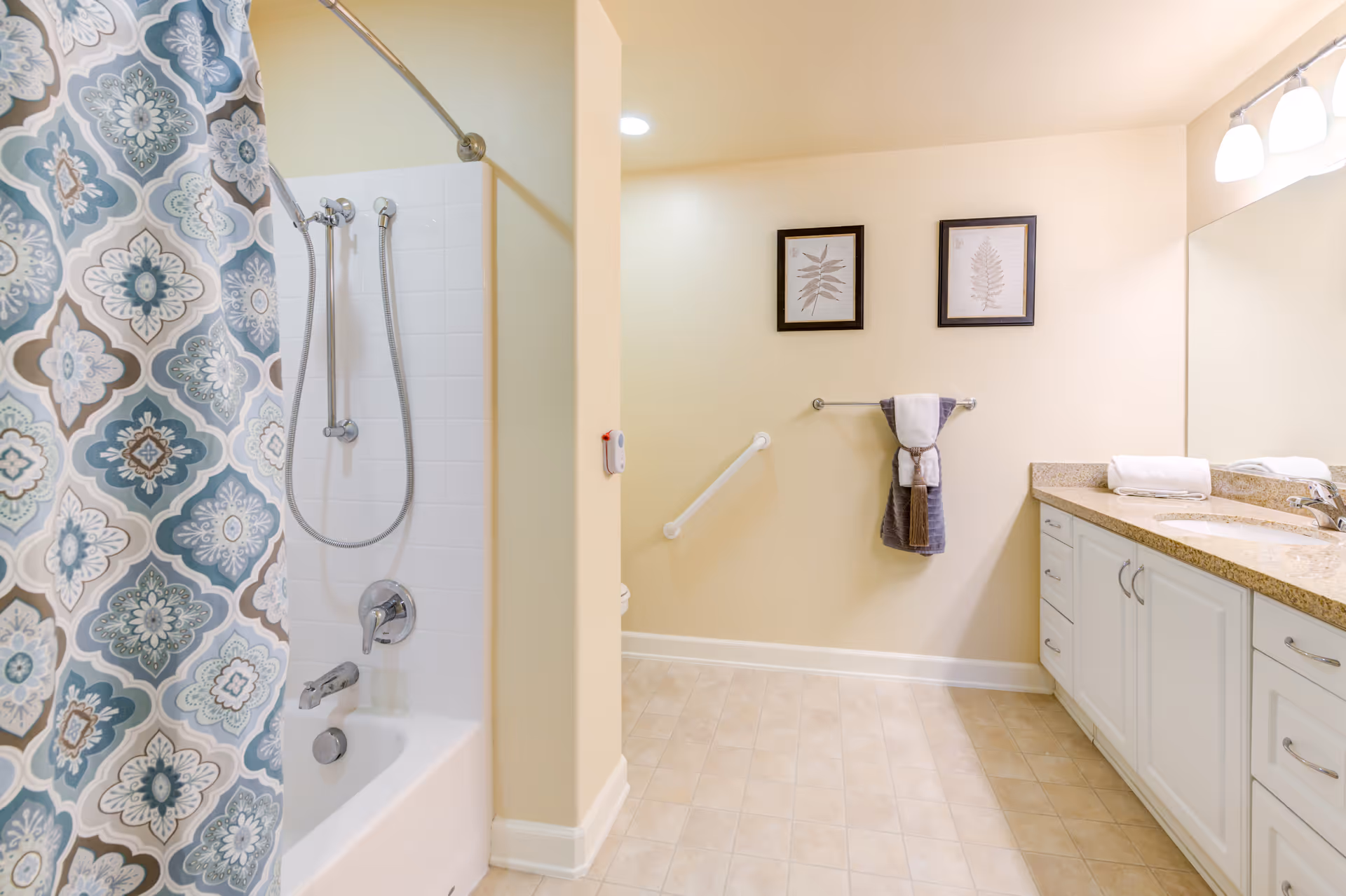 A clean and spacious bathroom with a bathtub and shower combination on the left, featuring a patterned blue and beige shower curtain. The bathroom has beige walls and tiled flooring. On the right side, there is a long countertop with a sink, white cabinets underneath, and a large mirror above. Two framed botanical prints hang on the wall above a towel rack holding folded towels. A grab bar is mounted on the wall near the towel rack.