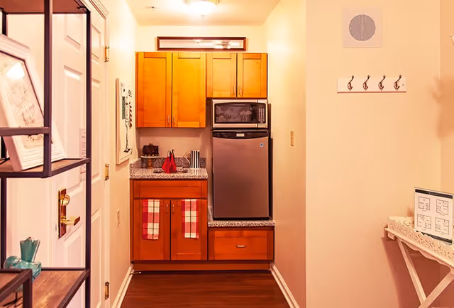 Small kitchenette area with wooden cabinets, a granite countertop, a stainless steel mini refrigerator, and a microwave on top. Two red and white checkered towels hang from the cabinet handles. There is a small sink with a red dish soap bottle and a few utensils. On the right wall, there is a coat rack with four hooks and a small table with a framed floor plan. The floor is dark wood, and the walls are painted light beige.