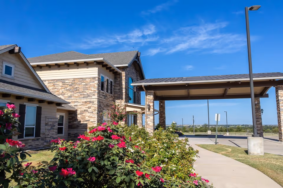 Exterior view of a senior living facility with stone and beige siding, a covered entrance supported by stone pillars, a sidewalk, and blooming pink flowers in the foreground under a clear blue sky.