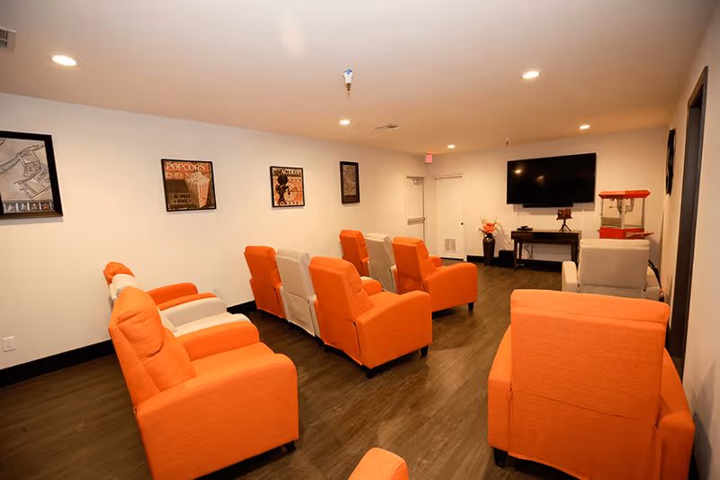 A small entertainment room with rows of orange and beige recliner chairs facing a wall-mounted TV and a popcorn machine.