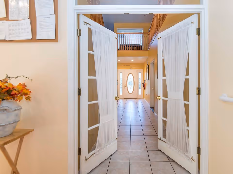View through open double doors with white curtains into a hallway with tiled floor leading to a front door with an oval glass window. The hallway has beige walls, a bulletin board on the left wall, and a wooden railing on the upper level.