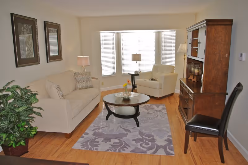 A cozy living room with a beige sofa on the left, a white armchair near a bay window with blinds, a round wooden coffee table on a patterned rug, a wooden cabinet on the right, a black chair, two framed pictures on the wall, and a potted plant in the corner.