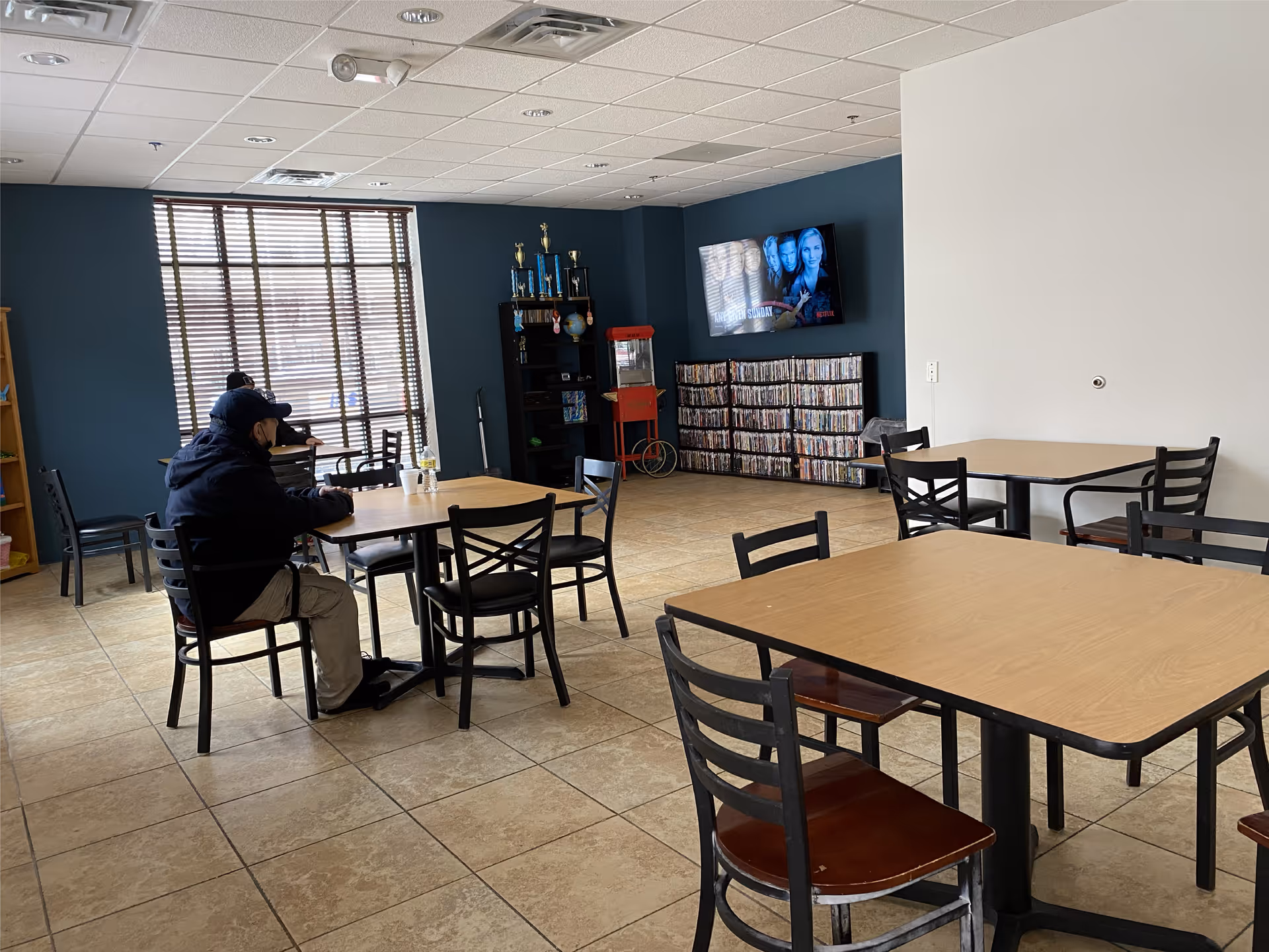 A common area with several tables and chairs, two people sitting at one table near a window with blinds. A TV mounted on a blue wall displays a show, with shelves below holding DVDs and a popcorn machine nearby. The floor is tiled and the room has a mix of blue and white walls.