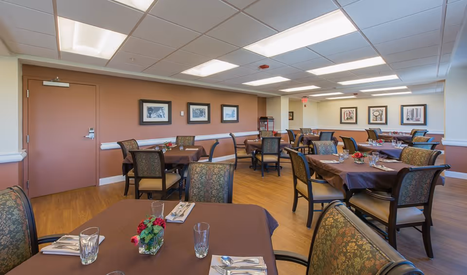 A dining room in a senior living facility with several tables covered in brown tablecloths, each set with glasses, napkins, and silverware. The chairs have patterned upholstery, and the walls are decorated with framed artwork. The room has a wooden floor and a ceiling with fluorescent lighting.