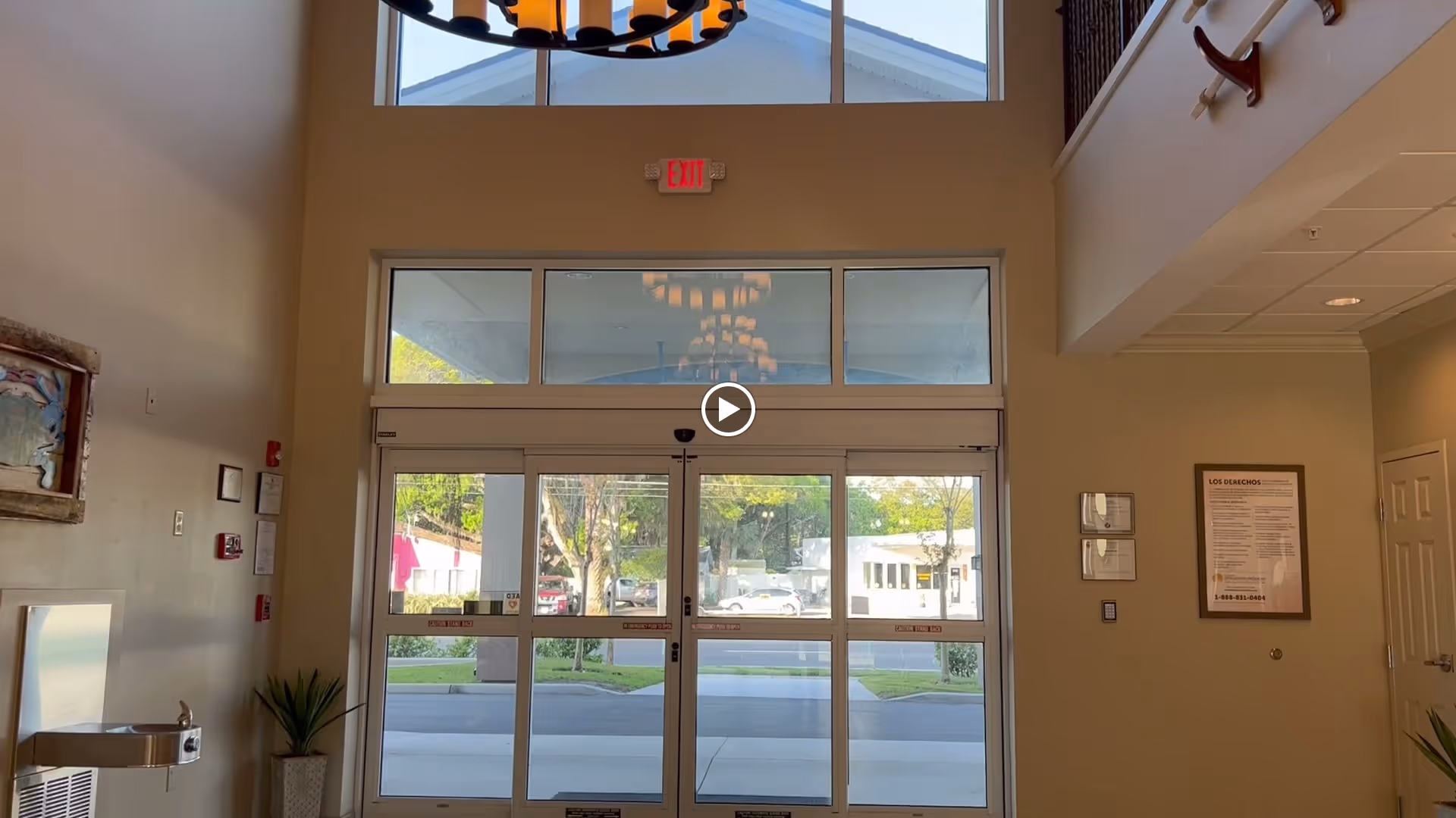 Interior view of the entrance lobby of a senior living facility with large glass double doors, a chandelier hanging from the ceiling, an exit sign above the doors, a water fountain on the left, and framed notices on the walls.