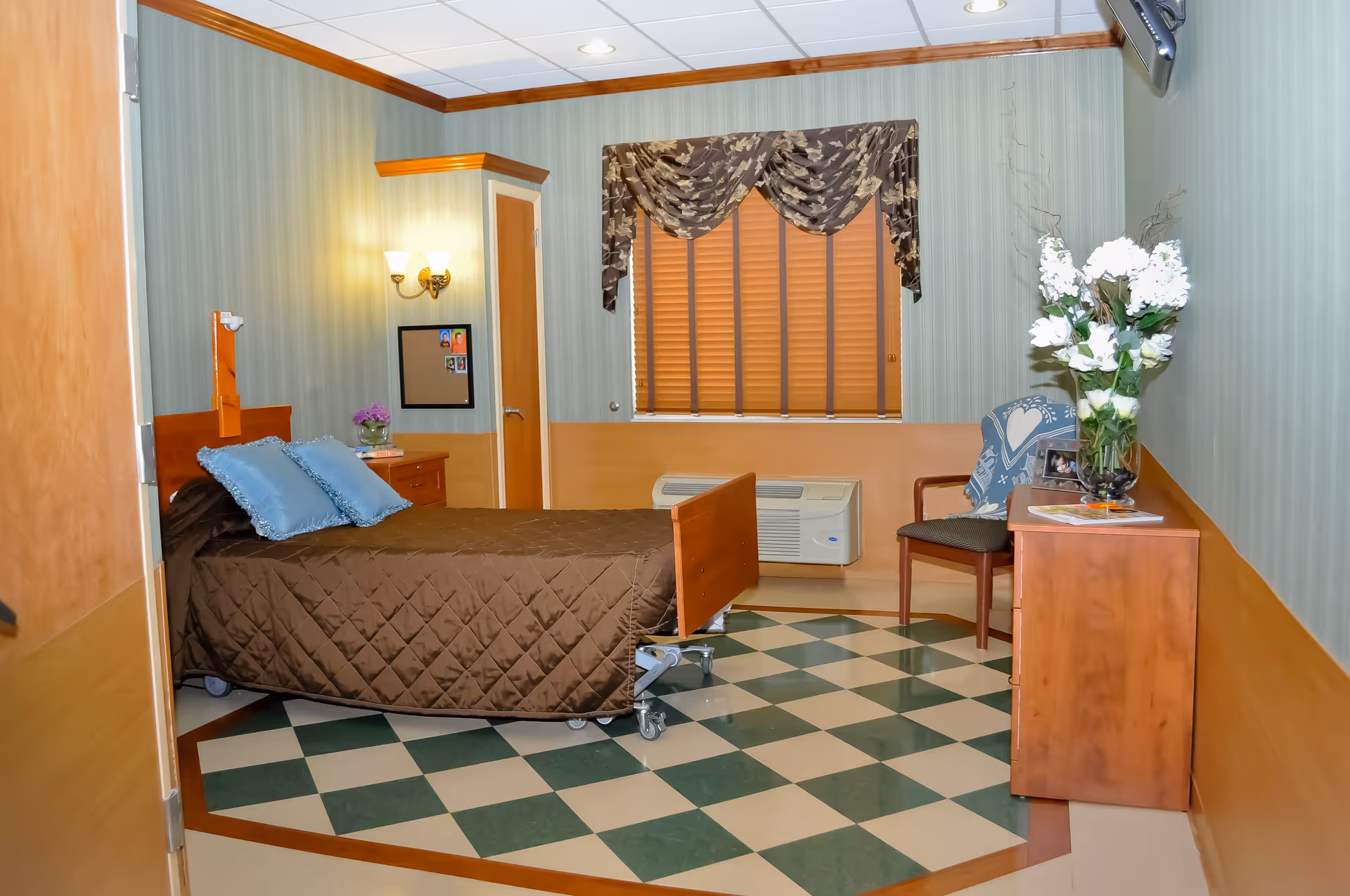 A neatly arranged bedroom in a senior living facility with a single bed covered in a brown quilt and two blue pillows. The room has green and beige checkered flooring, green striped wallpaper with wooden paneling on the lower half of the walls, a window with closed blinds and a decorative valance, a wooden dresser with a vase of white flowers, a chair with a blue blanket draped over it, and a wall-mounted air conditioning unit.