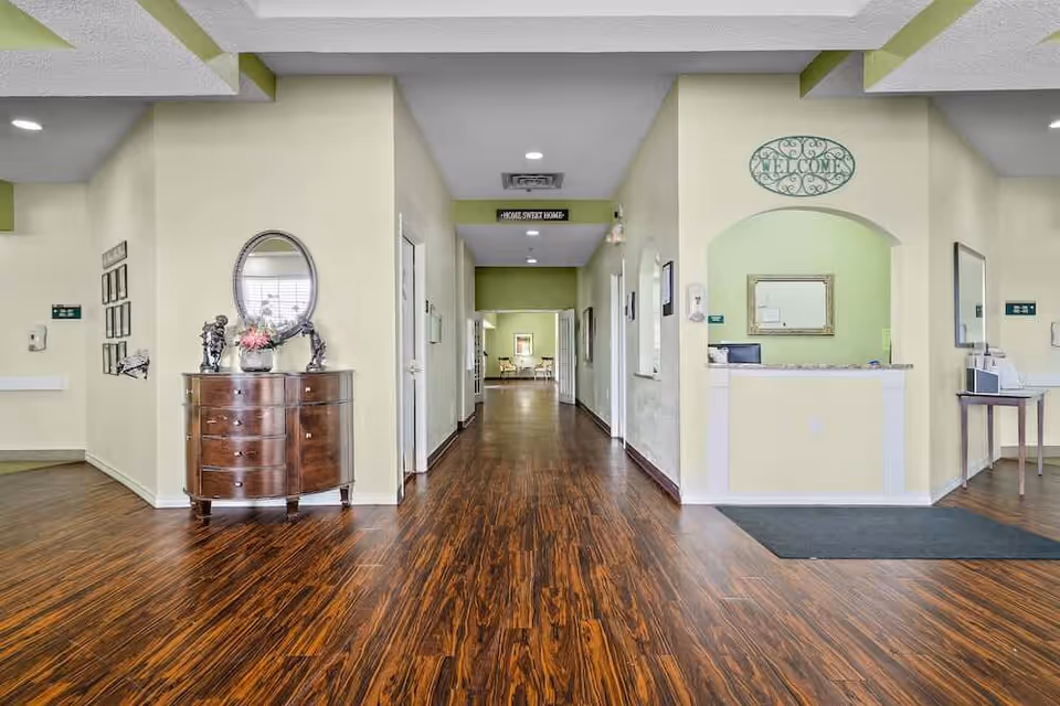 Interior view of a senior living facility hallway with wooden flooring, light yellow walls, and green accents. On the left, there is a wooden chest with decorative items and a round mirror above it. On the right, there is a reception desk area with a 'Welcome' sign above an arched opening. The hallway extends to a seating area visible in the distance.