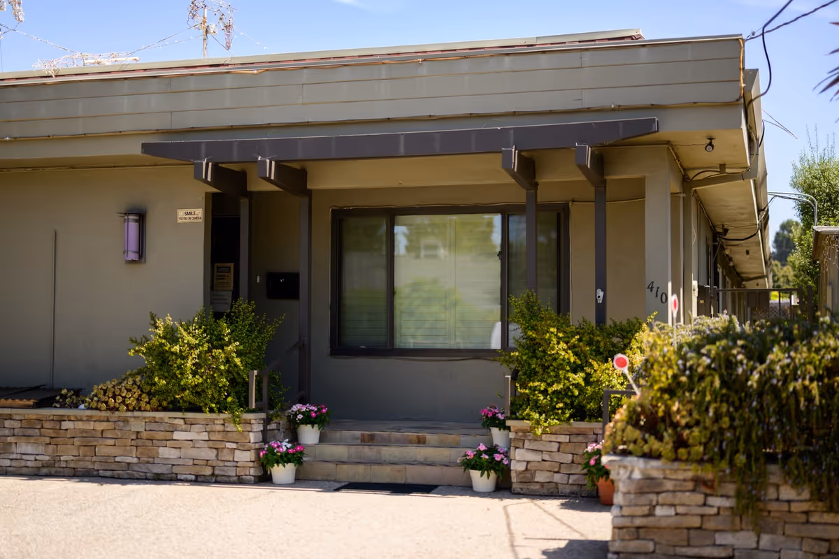 The front exterior of Maple House, a senior living facility, with a covered entrance, large window, and potted plants along the steps.