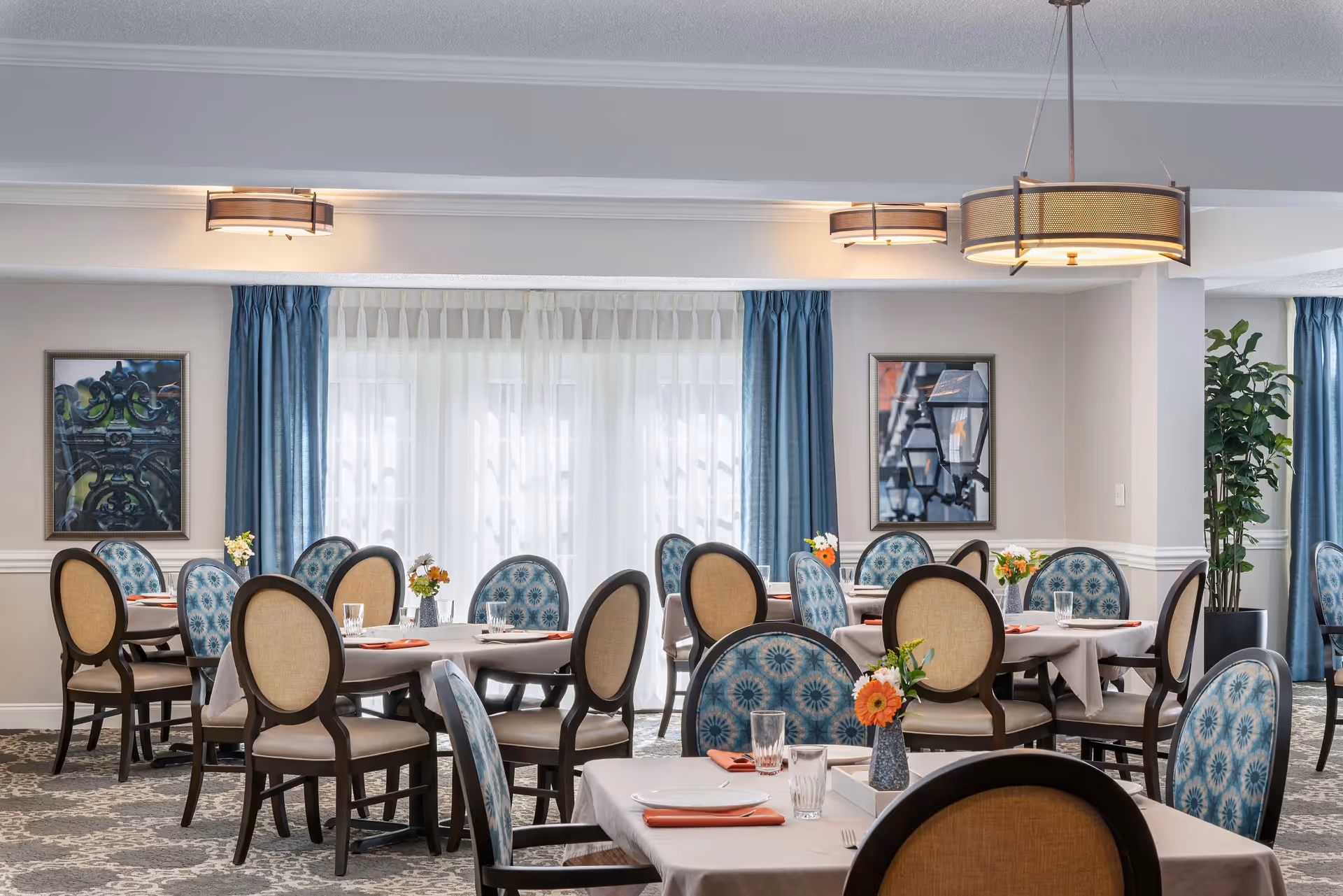 Dining room with round tables, upholstered chairs, place settings and floral centerpieces under pendant lights.