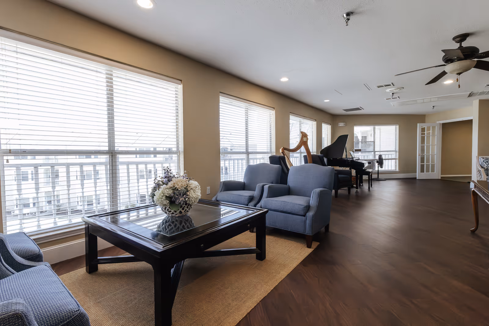 A bright and spacious senior living common area with large windows covered by white blinds. The room features comfortable blue armchairs around a dark wooden coffee table with a floral centerpiece. In the background, there is a grand piano and a harp, with ceiling fans overhead and wooden flooring throughout.