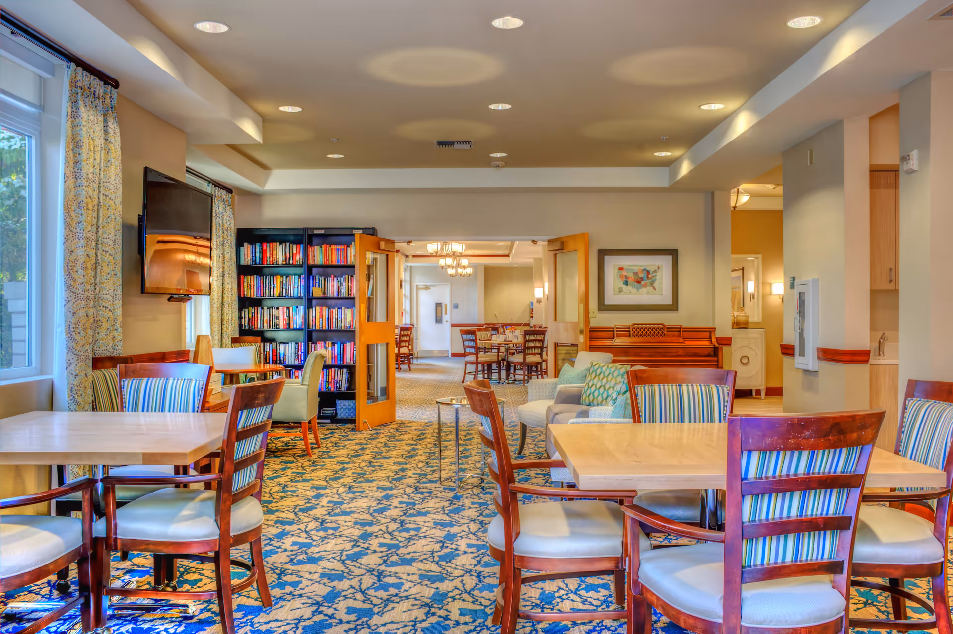 A bright and spacious common area in a senior living facility featuring several wooden tables with cushioned chairs arranged around them. There is a bookshelf filled with books against the far wall, a wall-mounted flat-screen TV, and large windows with patterned curtains letting in natural light. The room has a patterned carpet and recessed ceiling lights. In the background, there is another room with more tables and chairs, and a framed map on the wall.