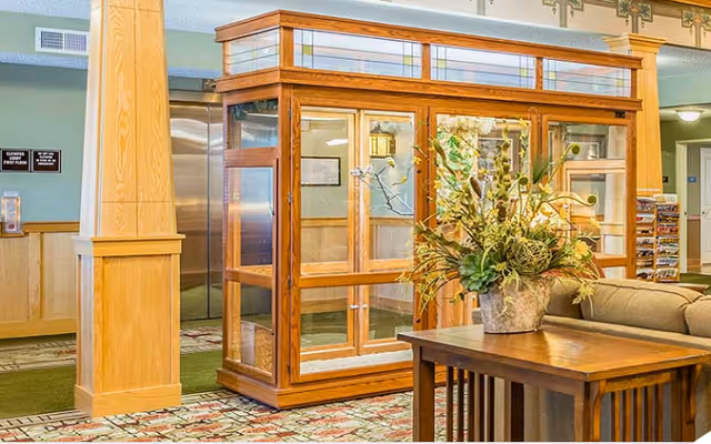 Interior view of a senior living facility lobby area featuring a wooden display case with glass panels, a wooden table with a large floral arrangement, a beige couch, and an elevator in the background. The walls have wood paneling and light green paint, with a patterned carpet on the floor.