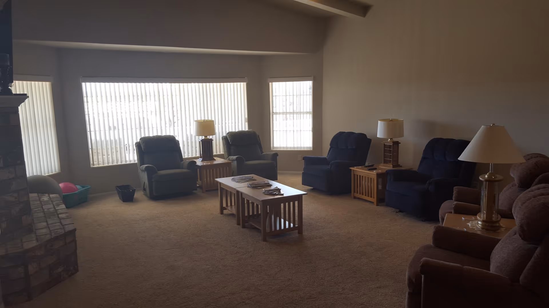 A senior living facility common room with several recliner chairs arranged around a wooden coffee table. The room has beige carpet, large windows with vertical blinds, and multiple table lamps on wooden side tables. There is a brick fireplace on the left side.