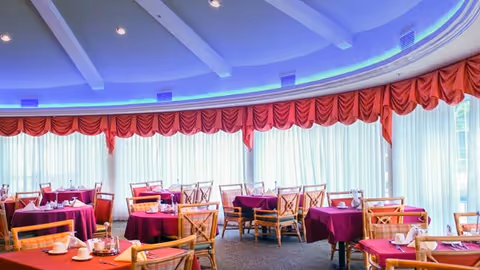 Bright circular dining room with round tables covered in purple tablecloths, wooden chairs, and a domed ceiling with blue accent lighting and red drapes.