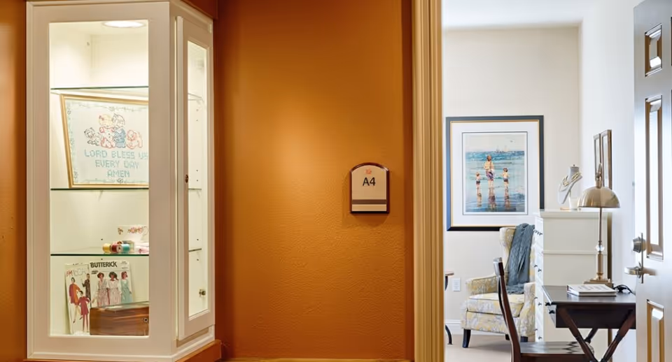 View of a hallway with a room labeled A4. On the left side, there is a white display cabinet with glass shelves containing decorative items including a framed cross-stitch, vintage magazines, and a wooden box. On the right side, through an open door, there is a cozy room with a patterned armchair, a framed picture of children playing at the beach on the wall, a white dresser, a desk with a lamp, and a chair.