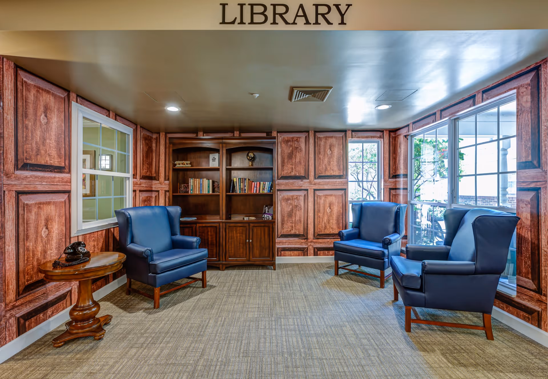 A cozy library room with wood-paneled walls and a carpeted floor. The room contains a wooden bookshelf filled with books, three blue upholstered armchairs arranged around the room, and a small round wooden side table with a vintage black rotary phone on it. Large windows allow natural light to enter the space.
