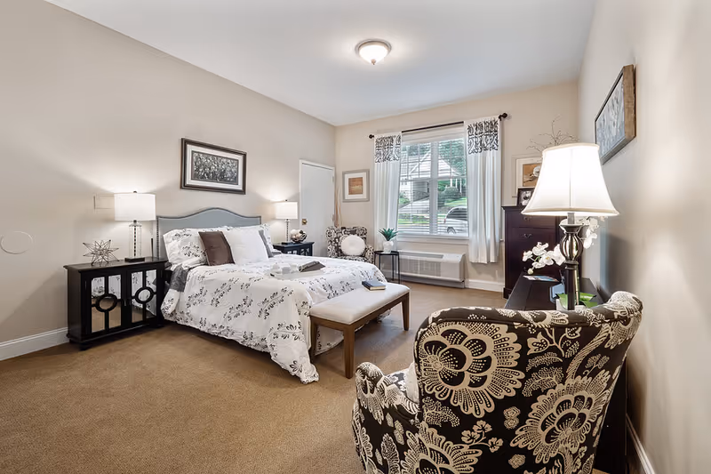 Well-lit furnished bedroom with a bed, nightstands, a patterned armchair, bench, and a window with curtains.