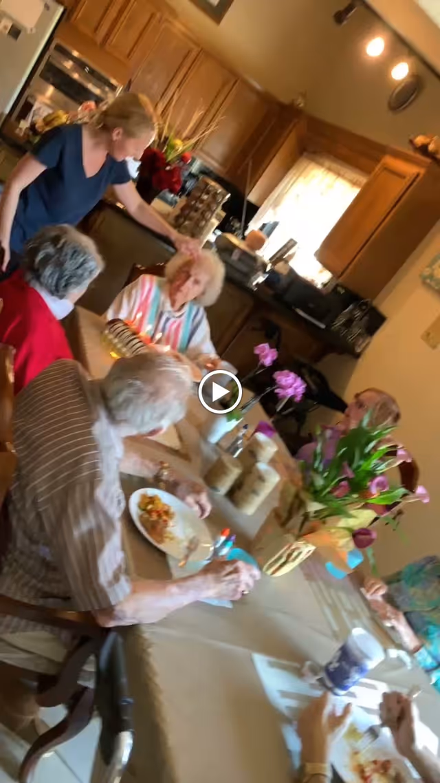 A group of elderly people seated around a dining table in a kitchen or dining area, with a caregiver standing and gently touching one elderly woman's head. The table is set with plates of food, cups, and decorative flowers. The room has wooden cabinets and a window with curtains letting in natural light.