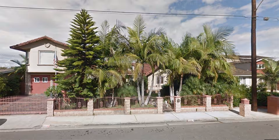 Exterior view of a residential-style building with a red gate and fence, surrounded by tall palm trees and other greenery, under a partly cloudy sky.