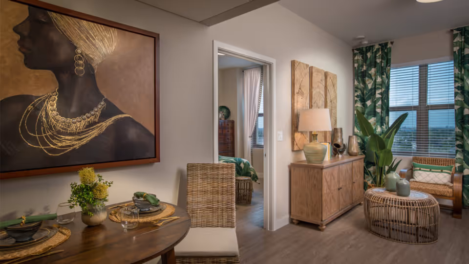 Interior view of a senior living facility showing a dining area with a round wooden table set for two, a wicker chair, and a large framed artwork of a woman with gold jewelry on the wall. Adjacent to the dining area is a living space with a wooden sideboard, decorative items, a lamp, a round wicker coffee table, a wicker armchair with cushions, and large windows with green leafy patterned curtains. A doorway leads to a bedroom with a bed and window visible.