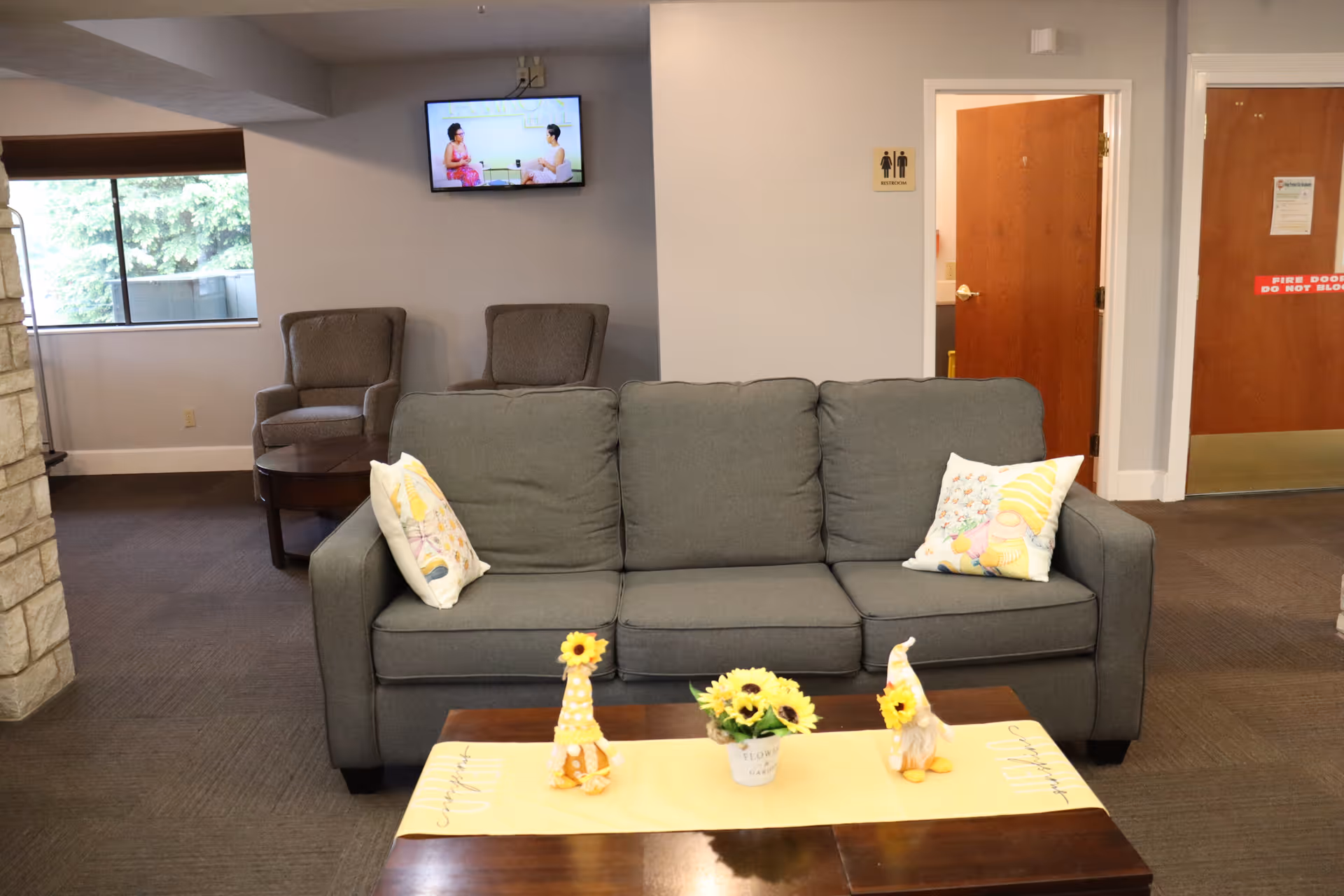 A cozy living room area with a gray sofa adorned with two decorative pillows. In front of the sofa is a wooden coffee table with a yellow runner and three small sunflower-themed decorations. Behind the sofa, there are two armchairs and a small round table near a window. A wall-mounted TV is showing a program with two people talking. There are two wooden doors on the right side, one with a restroom sign above it.