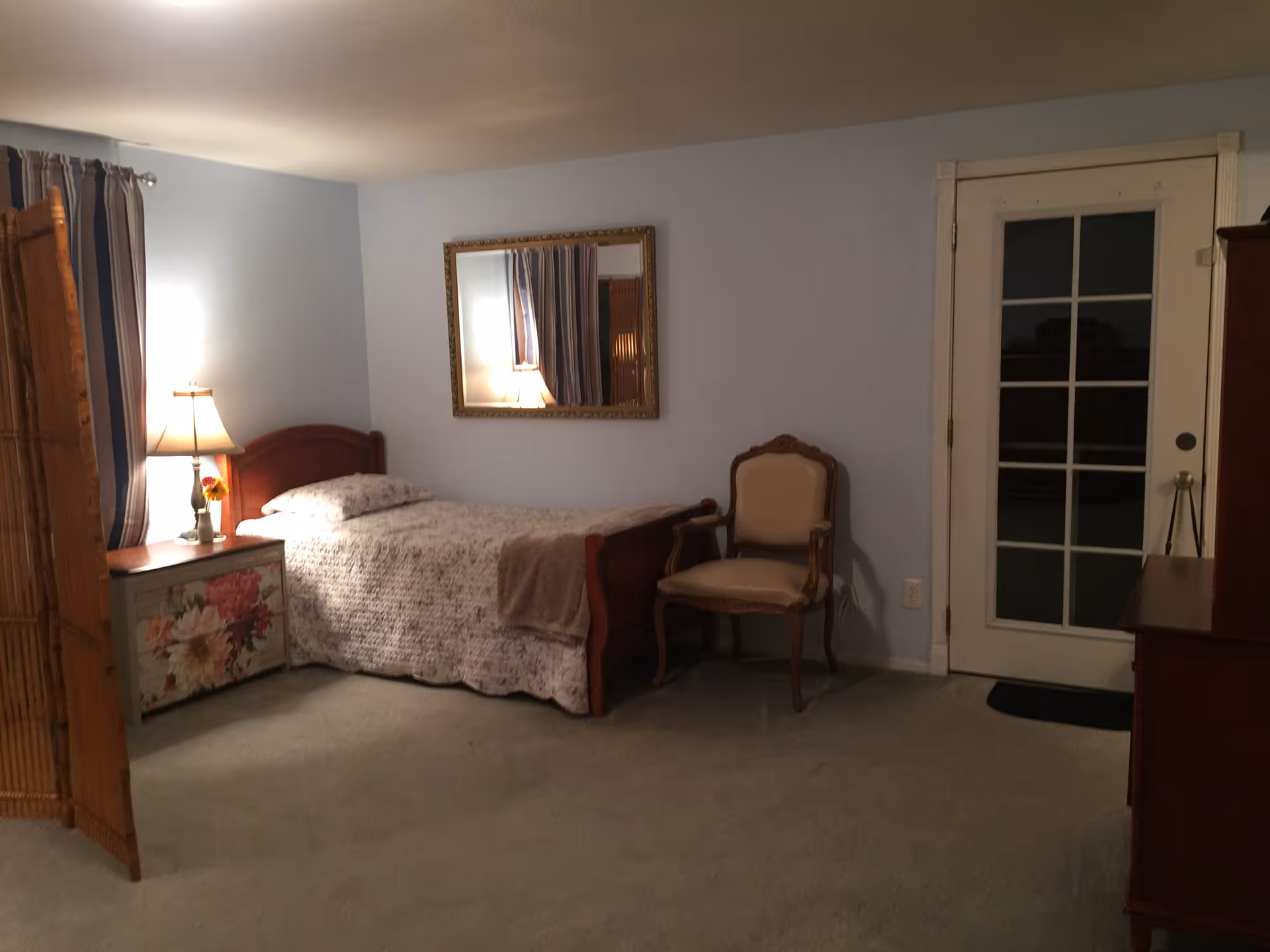 A cozy bedroom with a single bed covered in a floral quilt, a wooden headboard, and a bedside table with a lamp and a flower vase. There is a decorative mirror on the light blue wall above the bed, an upholstered armchair next to a glass-paneled door, and a wooden dresser on the right side. The room has carpeted flooring and soft lighting.
