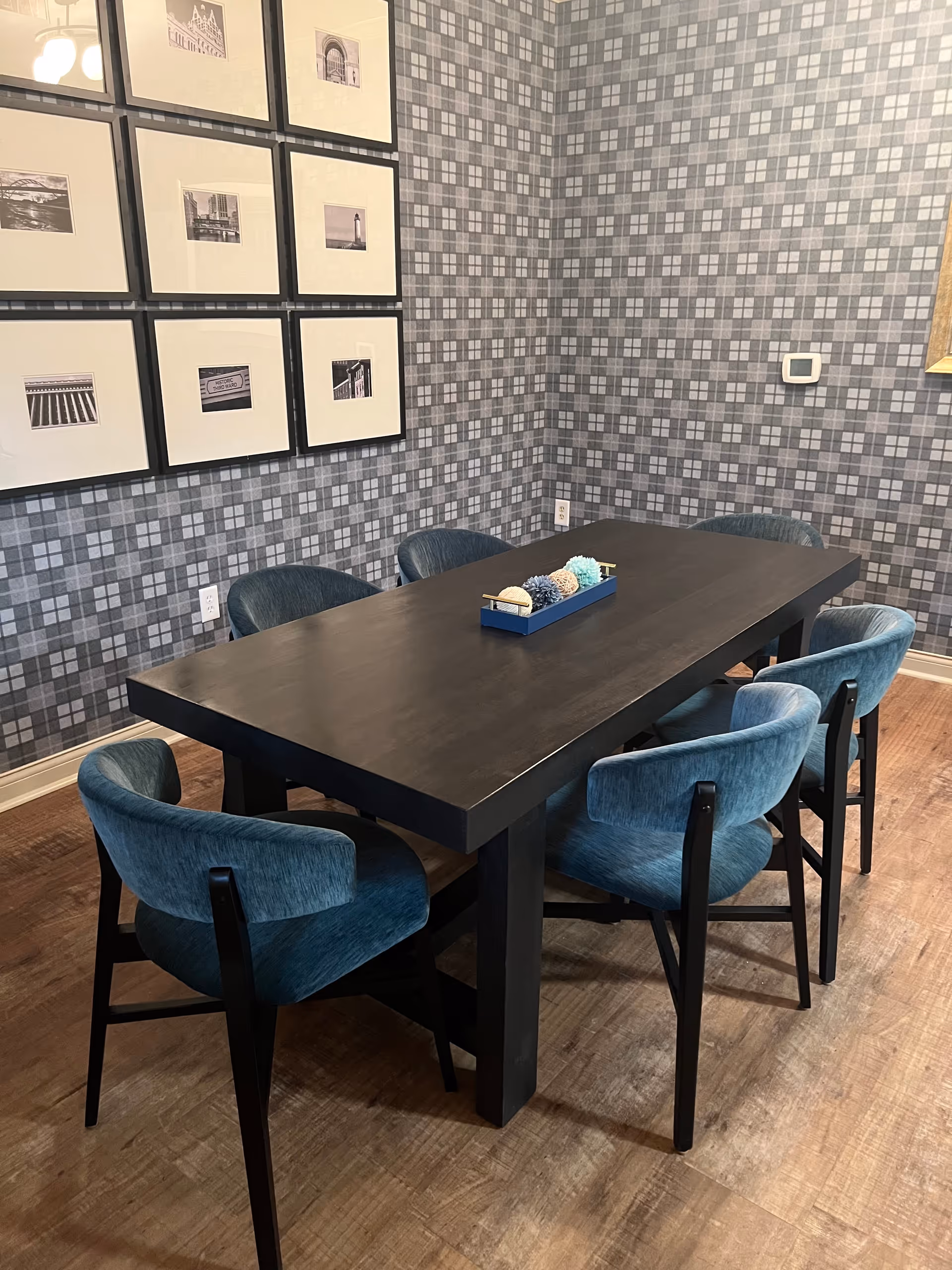 A modern dining area with a dark rectangular table surrounded by six blue upholstered chairs. The walls feature a gray and white checkered pattern wallpaper, and there are nine framed black and white photographs arranged in a grid on one wall. A decorative centerpiece with various textured balls is placed on the table.