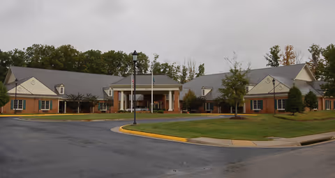 Exterior view of a single-story brick building with a gray roof, surrounded by a paved driveway and green lawn with trees in the background under an overcast sky.