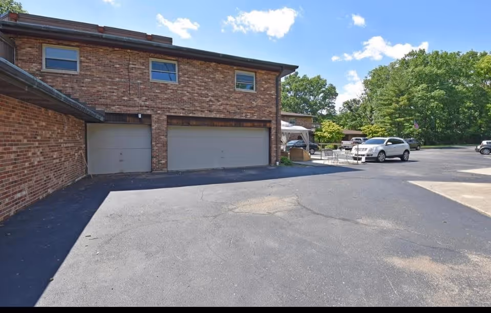 Exterior view of a brick building with three closed garage doors and a paved parking area in front. Several cars are parked near the building, and there are trees and greenery in the background under a partly cloudy blue sky.