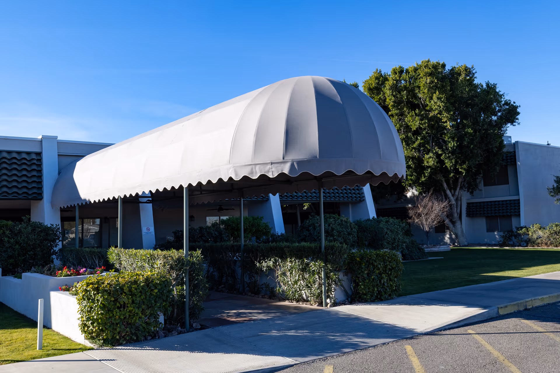 Entrance to a building with a large white canopy over the walkway, surrounded by green bushes and trees under a clear blue sky.