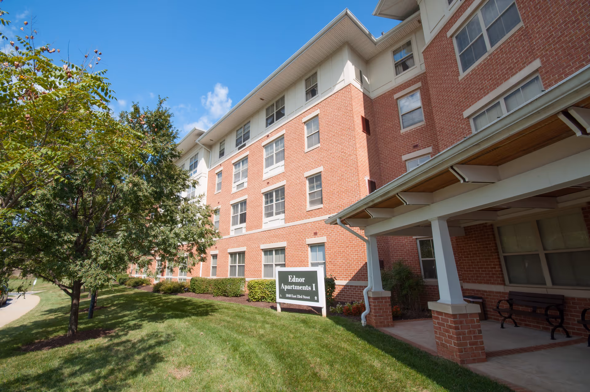 Exterior view of a multi-story brick apartment building named Ednor Apartments I with a green lawn, trees, and a covered entrance with benches under a clear blue sky.