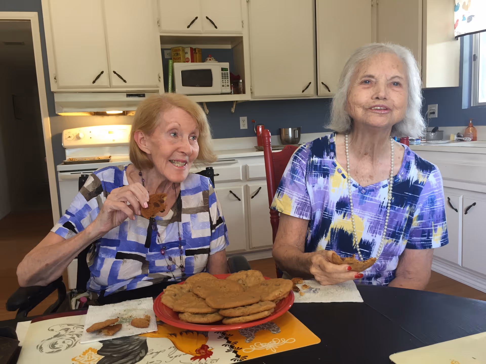 Two elderly women seated at a kitchen table smiling and eating cookies with a plate of cookies in front of them.