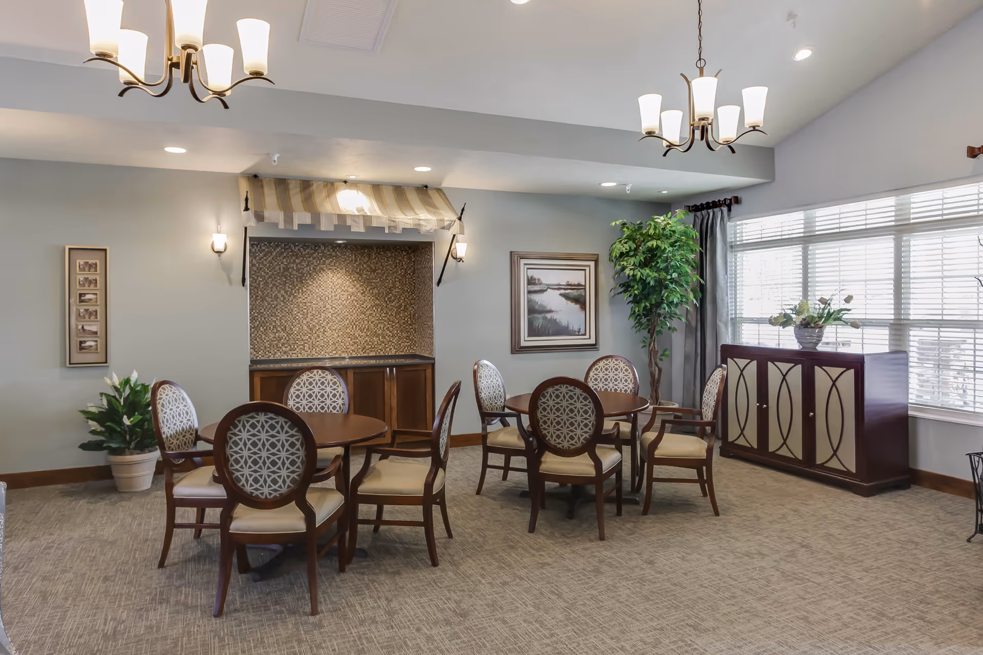 Bright communal dining area with round tables and patterned chairs, chandeliers, sideboard, plants, and large windows.