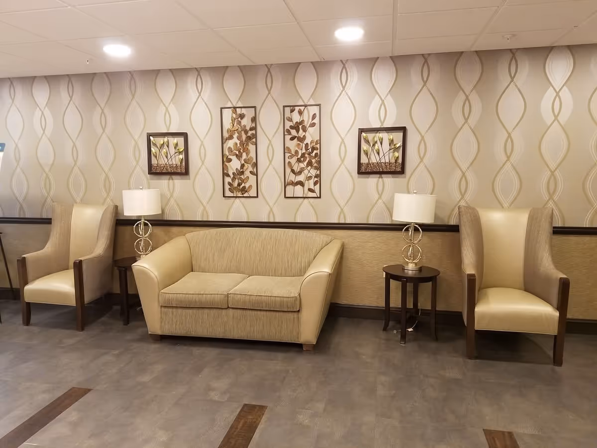 A seating area in a senior living facility with a beige loveseat sofa flanked by two beige armchairs. Each armchair has wooden armrests. There are two small round side tables with modern lamps on either side of the loveseat. The wall behind features patterned wallpaper with a wavy design and four framed metal leaf art pieces.