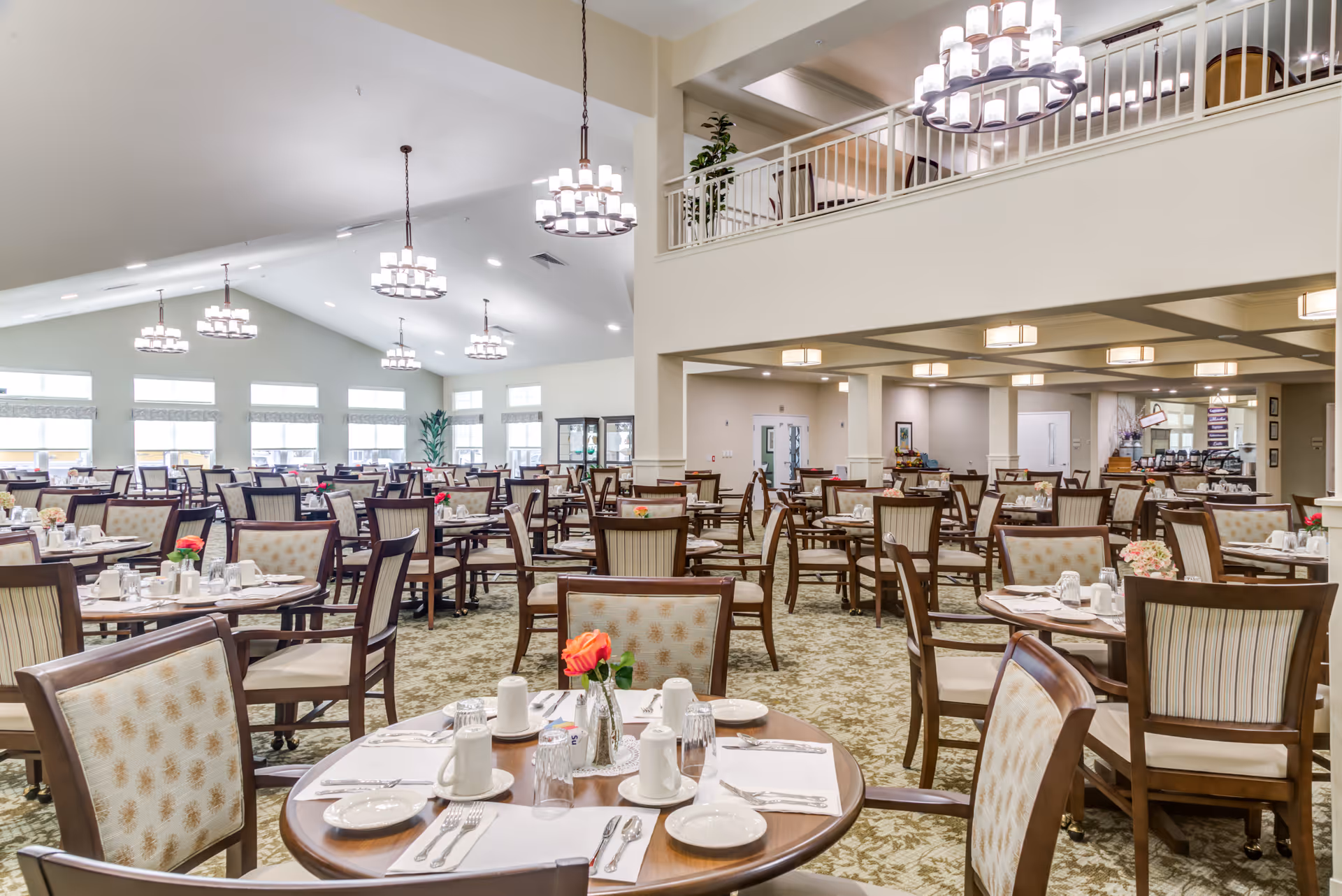 Spacious dining room in a senior living facility with multiple round tables set with white plates, cups, and silverware. Each table has a small vase with a red or pink flower. The room features high ceilings with multiple modern chandeliers, large windows letting in natural light, and a carpeted floor with a floral pattern. There is a mezzanine level with seating visible above.