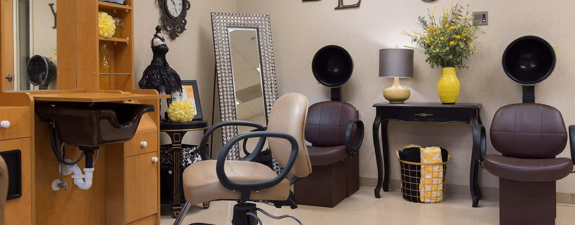Interior view of a salon area in a senior living facility featuring two brown salon chairs with hair dryers, a beige swivel chair, a wooden vanity with a black sink, a decorative black table with a yellow lamp and a yellow vase with flowers, a tall mirror with a jeweled frame, and various decorative items including a mannequin and framed pictures.