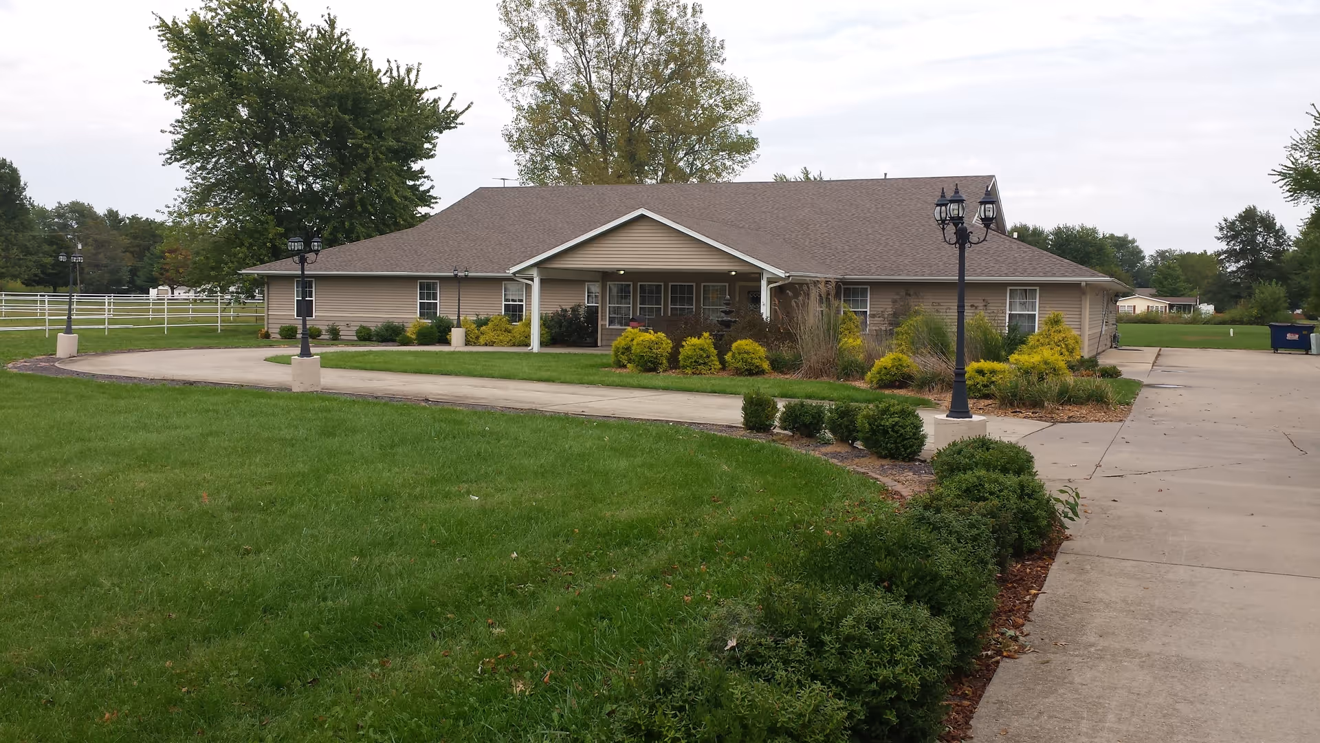 Single-story beige building with a covered entrance, surrounded by green grass, bushes, and trees. A curved concrete driveway leads to the entrance, with black lamp posts along the path. The sky is overcast.