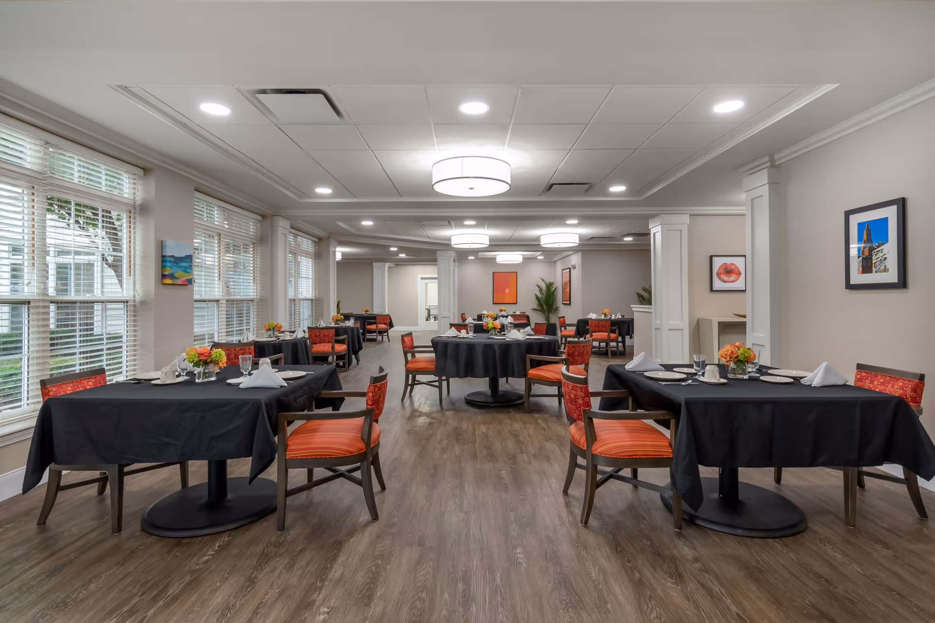 Bright dining room with round and rectangular tables set with black tablecloths, red upholstered chairs, and large windows.