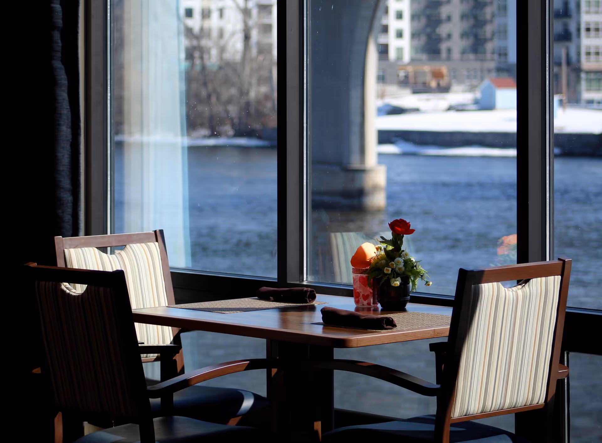 A dining table set for two with striped cushioned chairs is positioned next to large windows overlooking a river and a bridge. The table has placemats, rolled napkins, a small flower arrangement, and a container with napkins or utensils.