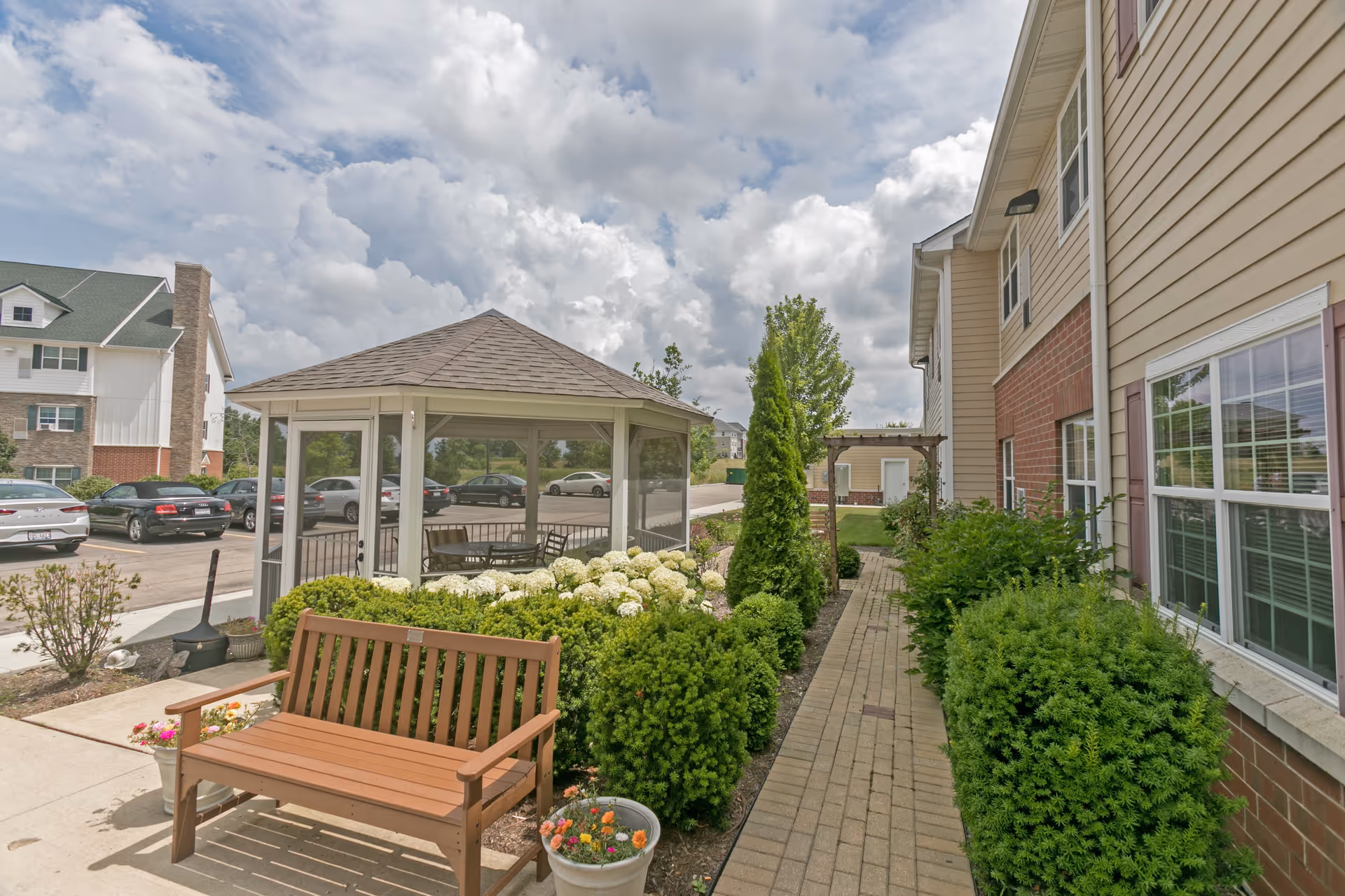 Outdoor area at Heritage Woods of Huntley featuring a wooden bench, potted flowers, a paved walkway lined with green bushes and white flowers, a gazebo with seating inside, and part of the building exterior with windows. Several parked cars and another building are visible in the background under a partly cloudy sky.