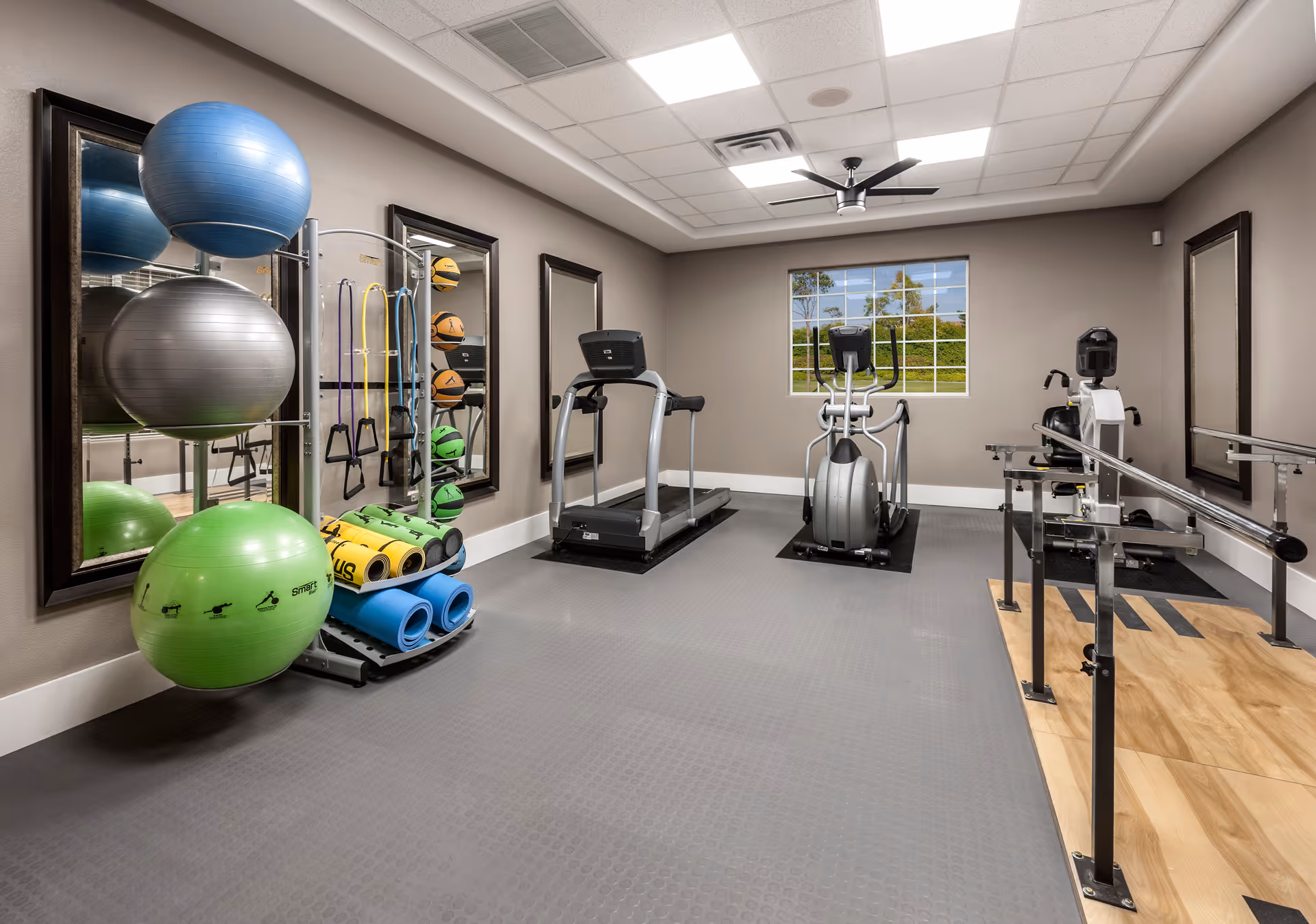 A fitness room with exercise equipment including a treadmill, elliptical machine, and a seated exercise machine with parallel bars. There are also large exercise balls, resistance bands, yoga mats, and medicine balls organized on racks against the wall. The room has gray flooring, beige walls, three large mirrors, a window showing greenery outside, and a ceiling fan.