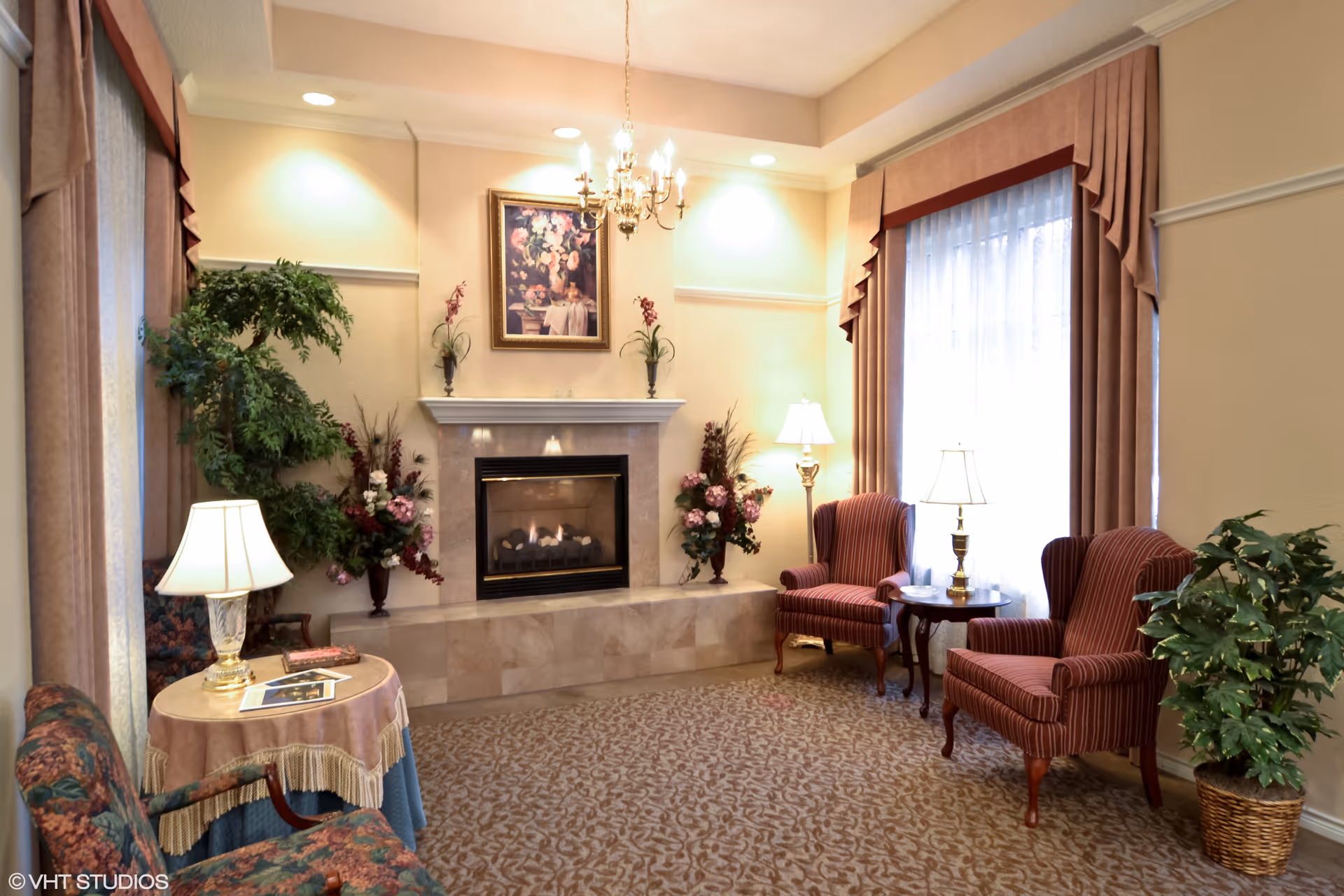 A cozy living room area with a lit fireplace in the center, flanked by floral arrangements. There are two red striped armchairs with a small round table and lamp between them on the right side, and a floral-patterned armchair with a round table and lamp on the left side. The room has beige walls, carpeted floor, large windows with sheer curtains and drapes, and a chandelier hanging from the ceiling.