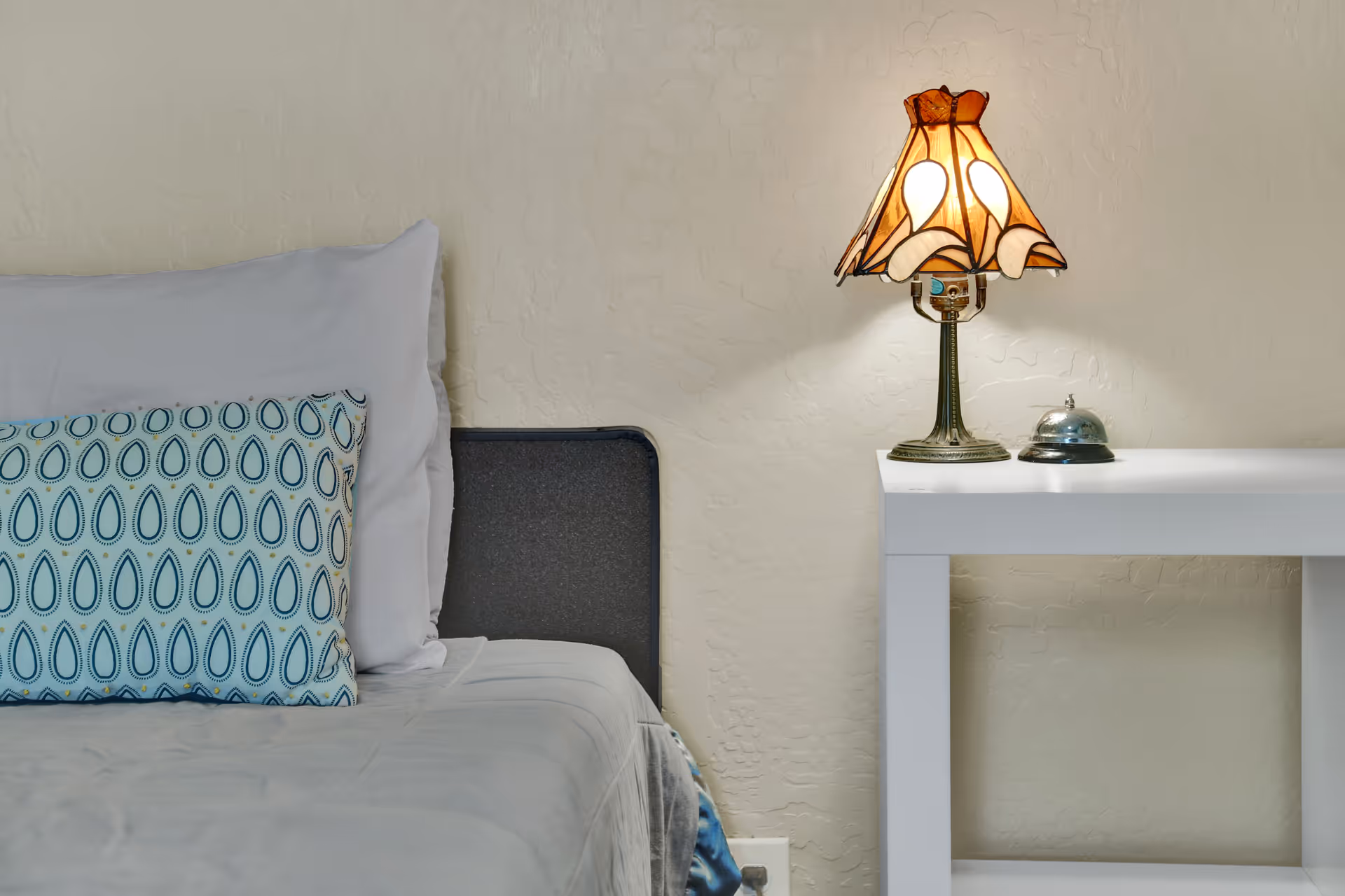 Side view of a neatly made bed with a patterned pillow beside a white nightstand holding a decorative lamp and a service bell.