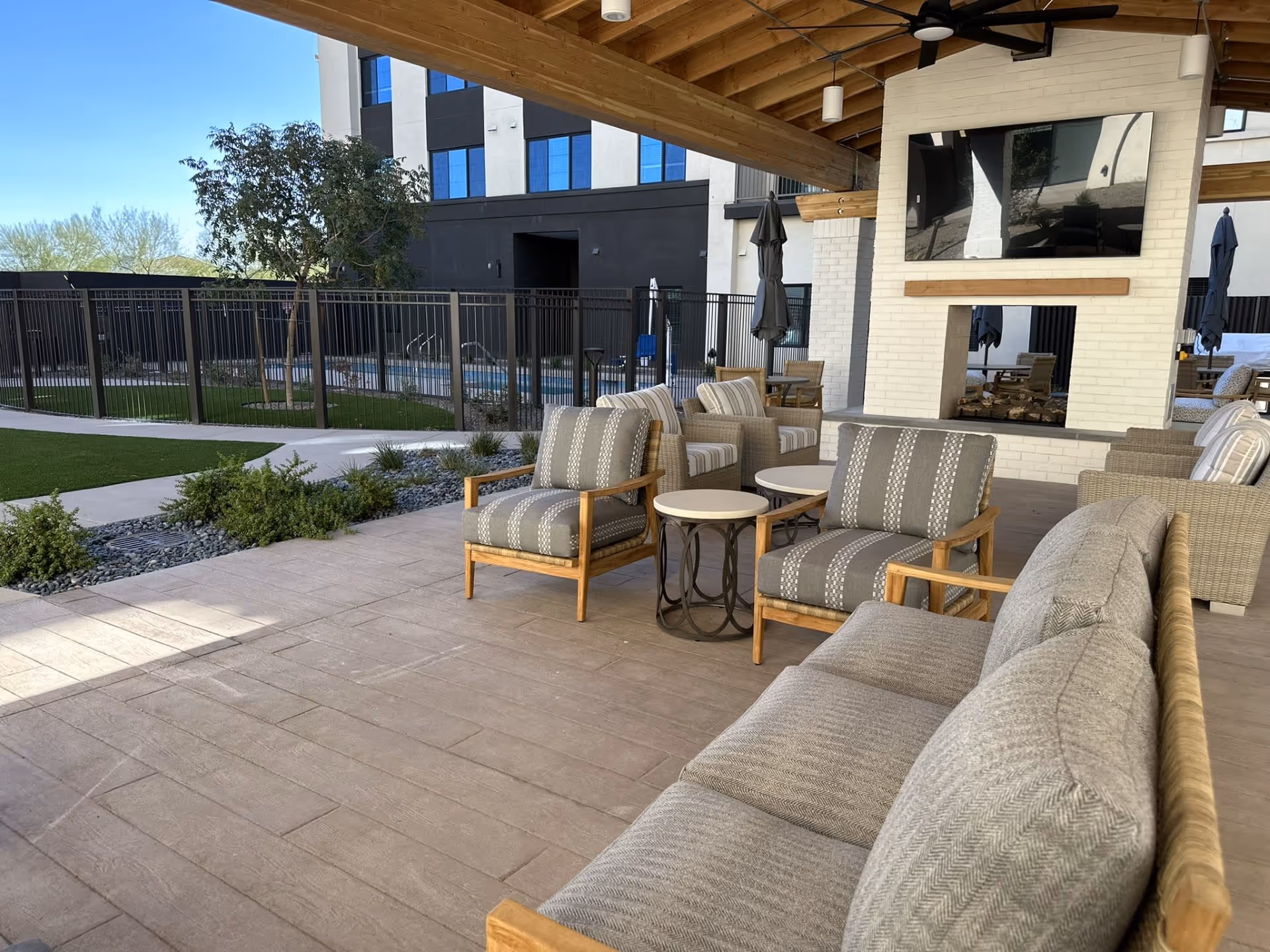 Outdoor covered patio area with cushioned seating including sofas and armchairs arranged around small tables. There is a white brick double-sided fireplace with a mounted flat-screen TV above it. In the background, there is a fenced pool area and a multi-story building with large windows.
